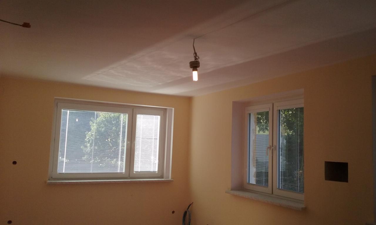 Pokój po szpachlowaniu z dwoma oknami w białych ramach i żarówką zwisającą z sufitu. Ściany w odcieniach beżu, przygotowane pod malowanie.