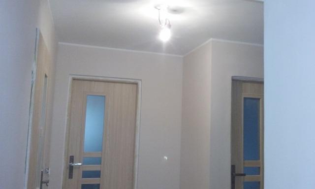 Jasne, świeżo odmalowane wnętrze z dwiema drewnianymi drzwiami z przeszkleniami, oświetlone centralnym punktowym światłem sufitowym.