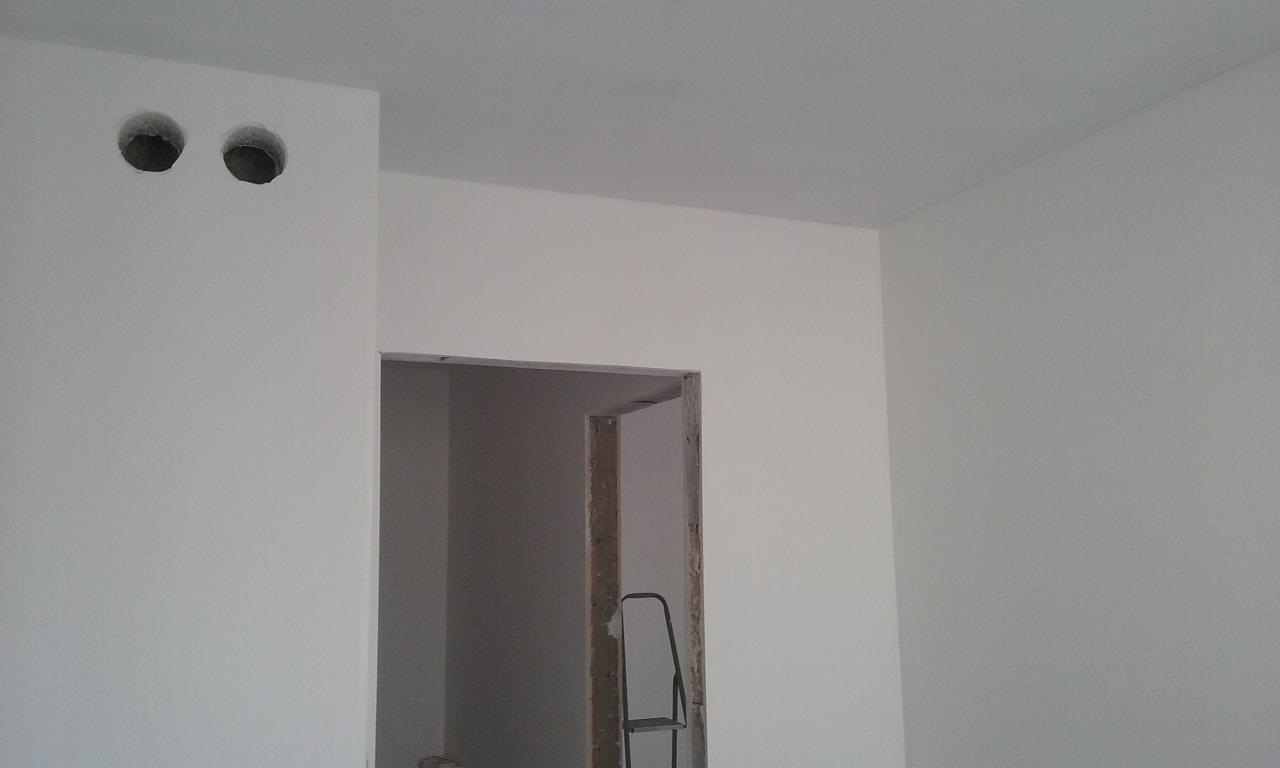 Białe ściany po szpachlowaniu, widoczne dwa otwory w ścianie i ościeżnica drzwi. W tle drabina. Jasne, naturalne światło.