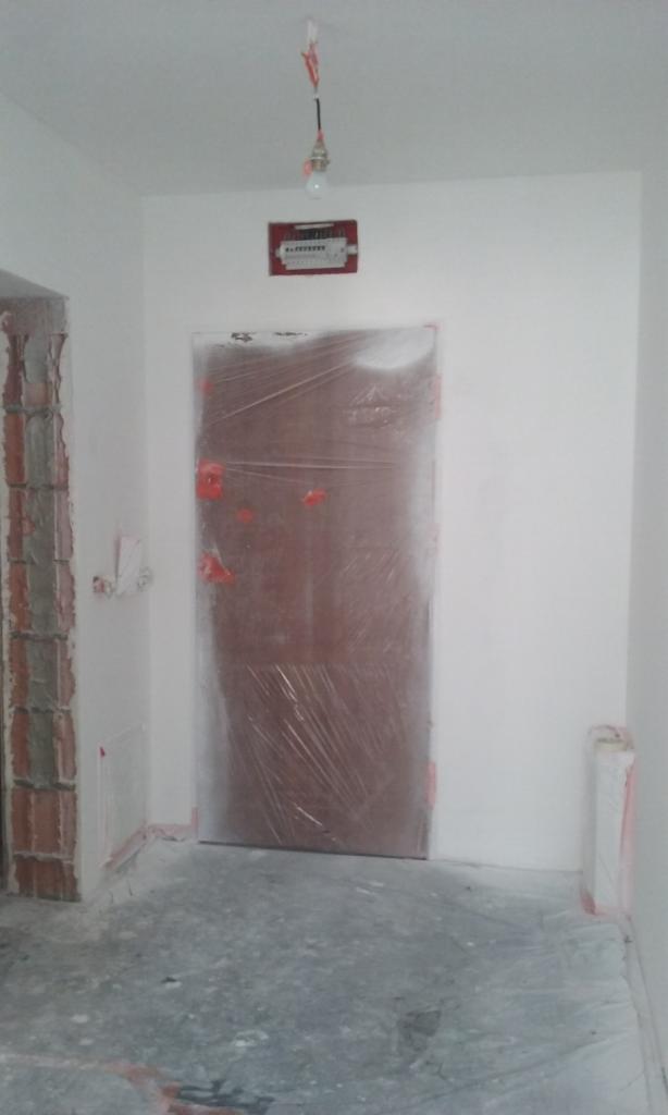 Przygotowanie do prac: zabezpieczone folią drzwi w pomieszczeniu z surowymi ścianami, widoczna instalacja elektryczna. Etap prac remontowych.