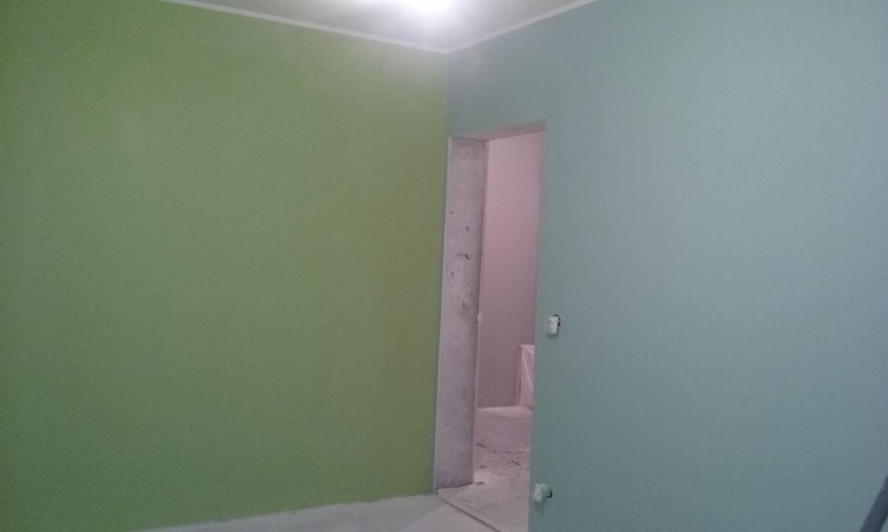 Ściana pomalowana na dwa kolory: zielony i niebieski, widoczne surowe obramowanie drzwi. W tle widoczny fragment innego pomieszczenia.