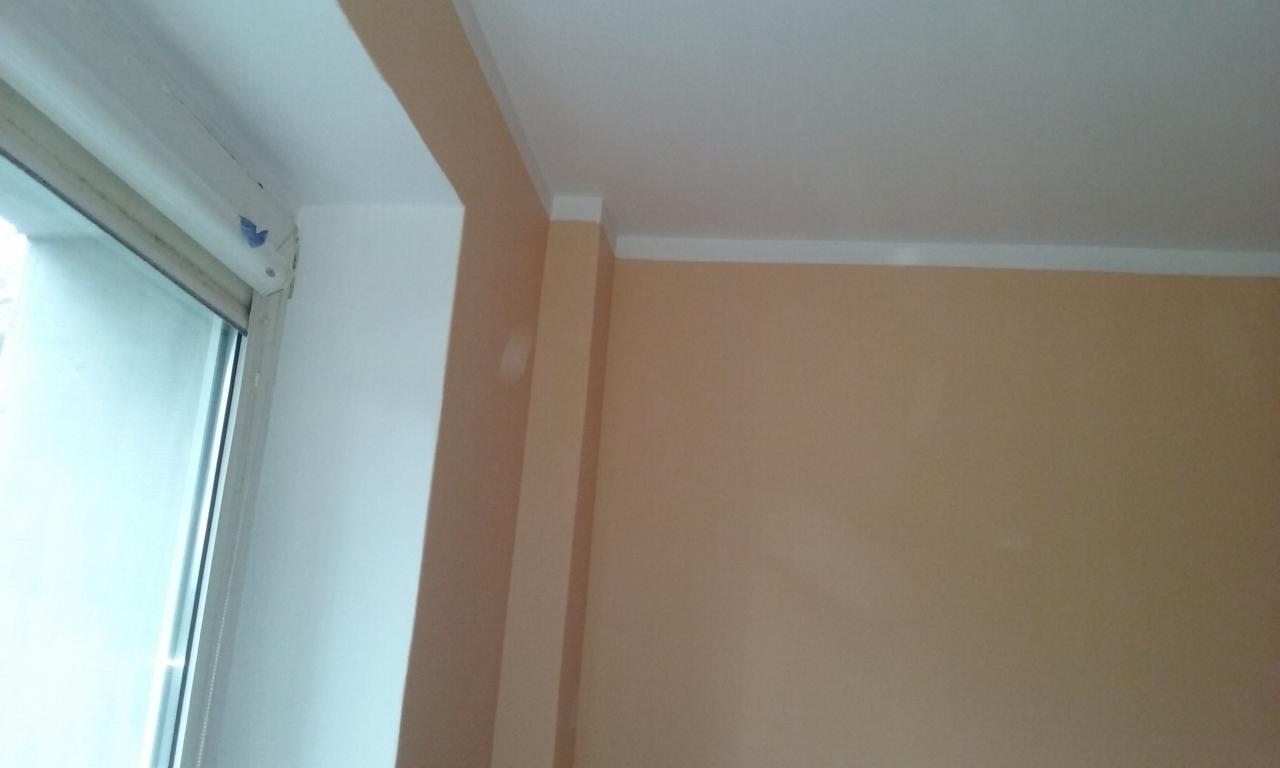 Ściana w odcieniach beżu i bieli po remoncie, widoczna listwa przypodłogowa i okno z roletą. Kąt ujęcia od dołu, z bliska, prezentuje detale wykończenia.