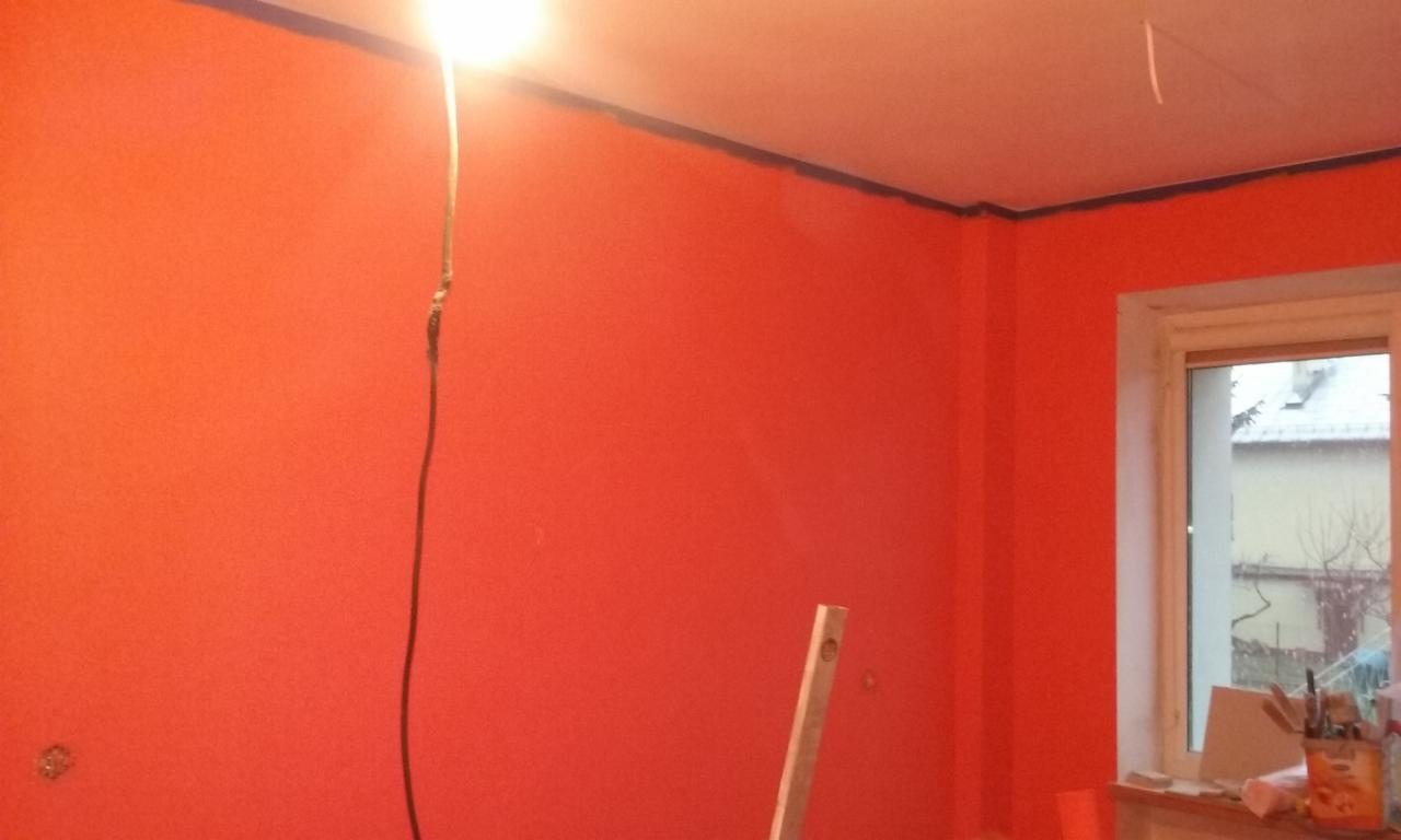 Ściana w intensywnym odcieniu czerwieni, zabezpieczona taśmą malarską przy suficie, z widoczną lampą i poziomnicą, w trakcie prac.