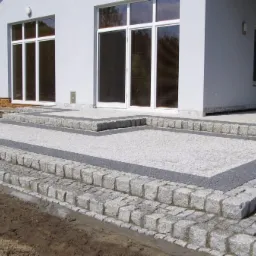 Nowoczesny taras z jasnoszarej kostki granitowej z ciemnoszarą obwódką, prowadzący do białego domu z dużymi oknami, widok z dołu.