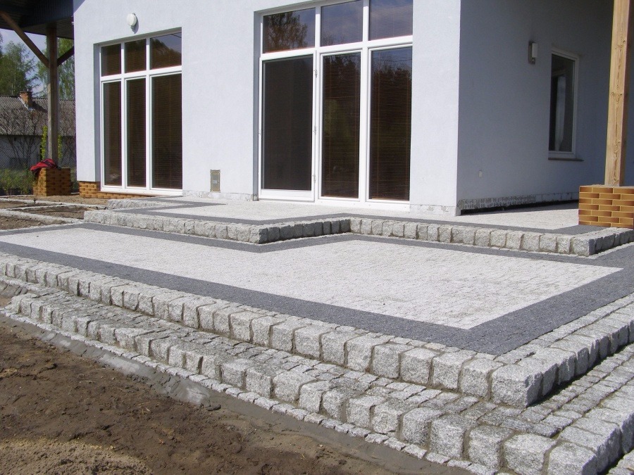 Nowoczesny taras z jasnoszarej kostki granitowej z ciemnoszarą obwódką, prowadzący do białego domu z dużymi oknami, widok z dołu.