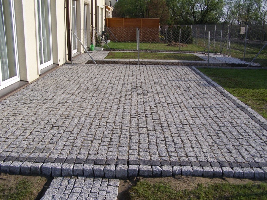 Nowo wyłożony taras z kostki granitowej w odcieniach szarości, z ciemniejszym obramowaniem, przylegający do jasnego budynku z dużymi oknami i widokiem na ogrodzenie z siatki oraz drewniany płot.