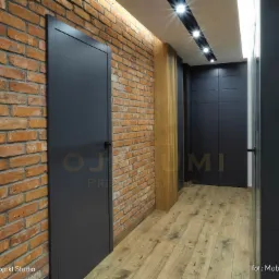 Wnętrze korytarza z ceglaną ścianą, drewnianą podłogą i ciemnymi drzwiami, oświetlone punktowymi lampami sufitowymi. Widoczny fragment logo firmy projektowej na drzwiach po lewej stronie.