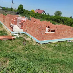 Wznoszone mury domu z czerwonej cegły ceramicznej, widoczne zbrojenia i paleta z cegłami na fundamencie, trawa na pierwszym planie, w tle dom z czerwonym dachem.
