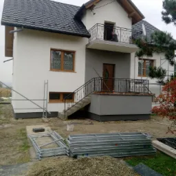 Dwupiętrowy dom z białą elewacją i ciemną dachówką, z widocznymi rusztowaniami i stertą żwiru na posesji, sugerujący prace wykończeniowe.