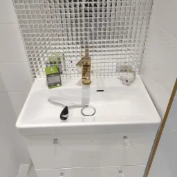 Biała umywalka z szafką, złotą baterią i mozaiką na ścianie, uzupełniona o akcesoria łazienkowe: ściągaczkę do wody i odświeżacz powietrza.