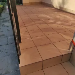 Jasnobrązowe płytki ceramiczne ułożone na tarasie z dwoma stopniami, widoczne fragmenty balustrady i elewacji budynku w ciepłym oświetleniu.