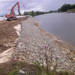 Brzeg zbiornika wodnego umocniony siatką gabionową wypełnioną kamieniami, w tle koparka i betonowe bariery, na pierwszym planie kwitnący oset.