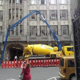 Żółta betonomieszarka KANN Beton z wysięgnikiem pompy SCHWING na tle budynku Passage, fotografowana przez kobietę zza ozdobnej bramy.