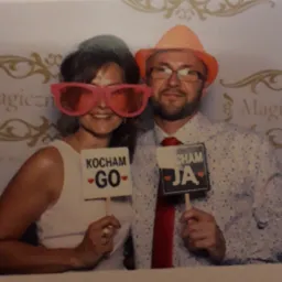 Para w śmiesznych okularach i kapeluszach pozuje z rekwizytami z napisem 'Kocham Go' i 'Kocham Ja' na tle dekoracyjnej ścianki z napisem 'Magiczne'