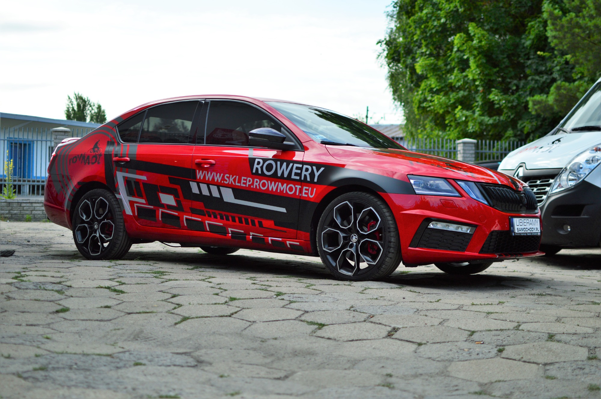 Czerwona Skoda Octavia z czarnymi felgami i grafiką reklamową na boku, z napisem 'ROWERY WWW.SKLEP.ROWMOT.EU', zaparkowana na kostce brukowej.