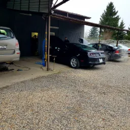 Warsztat samochodowy z kilkoma samochodami, w tym czarny Ford Mustang z otwartą maską, stojący na żwirowym parkingu przed budynkiem z otwartymi wrotami, gdzie widoczny jest mechanik.