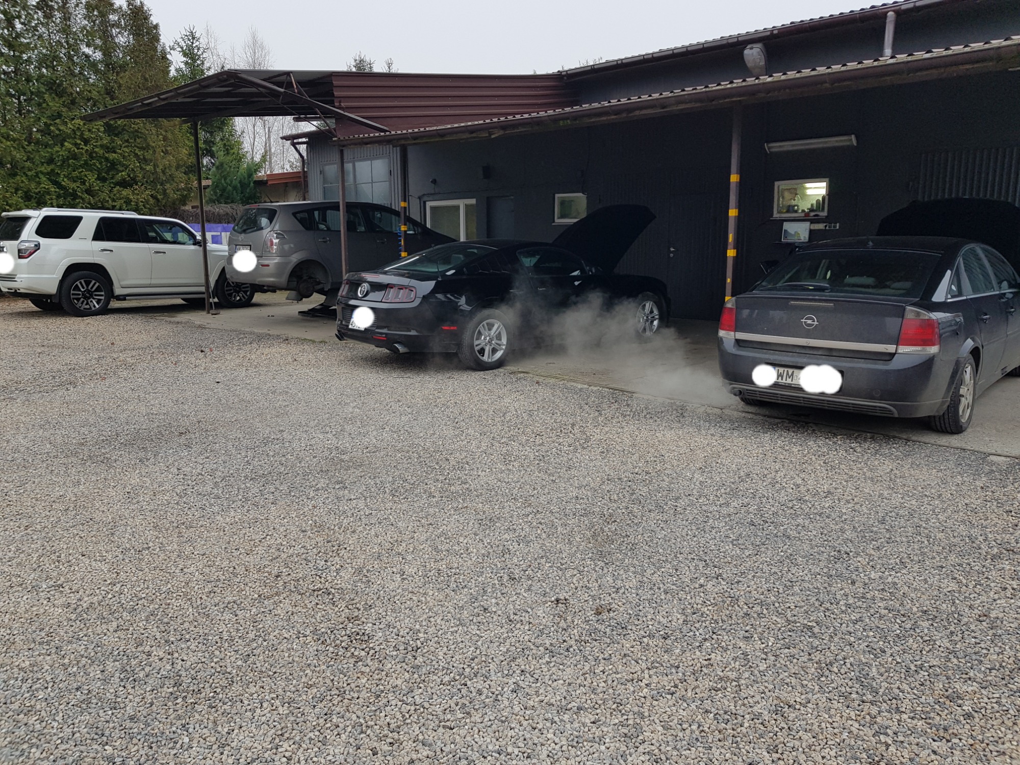 Czarny Ford Mustang z otwartą maską, wydobywający się dym, na tle warsztatu samochodowego z innymi pojazdami, w tym jeden na podnośniku.