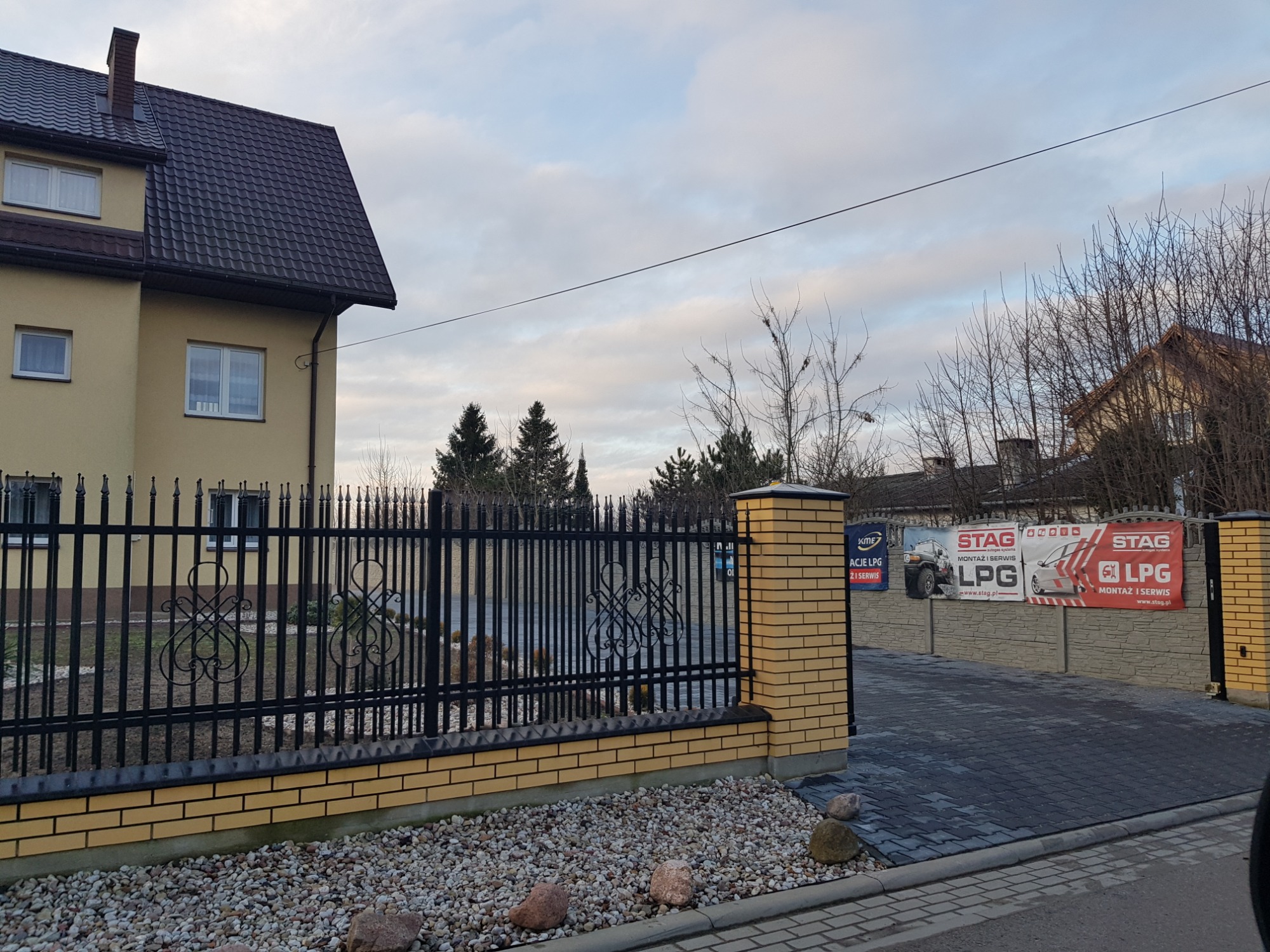 Budynek z banerami reklamowymi systemów montażu LPG widocznymi za ozdobnym, czarnym ogrodzeniem z ceglaną podmurówką.