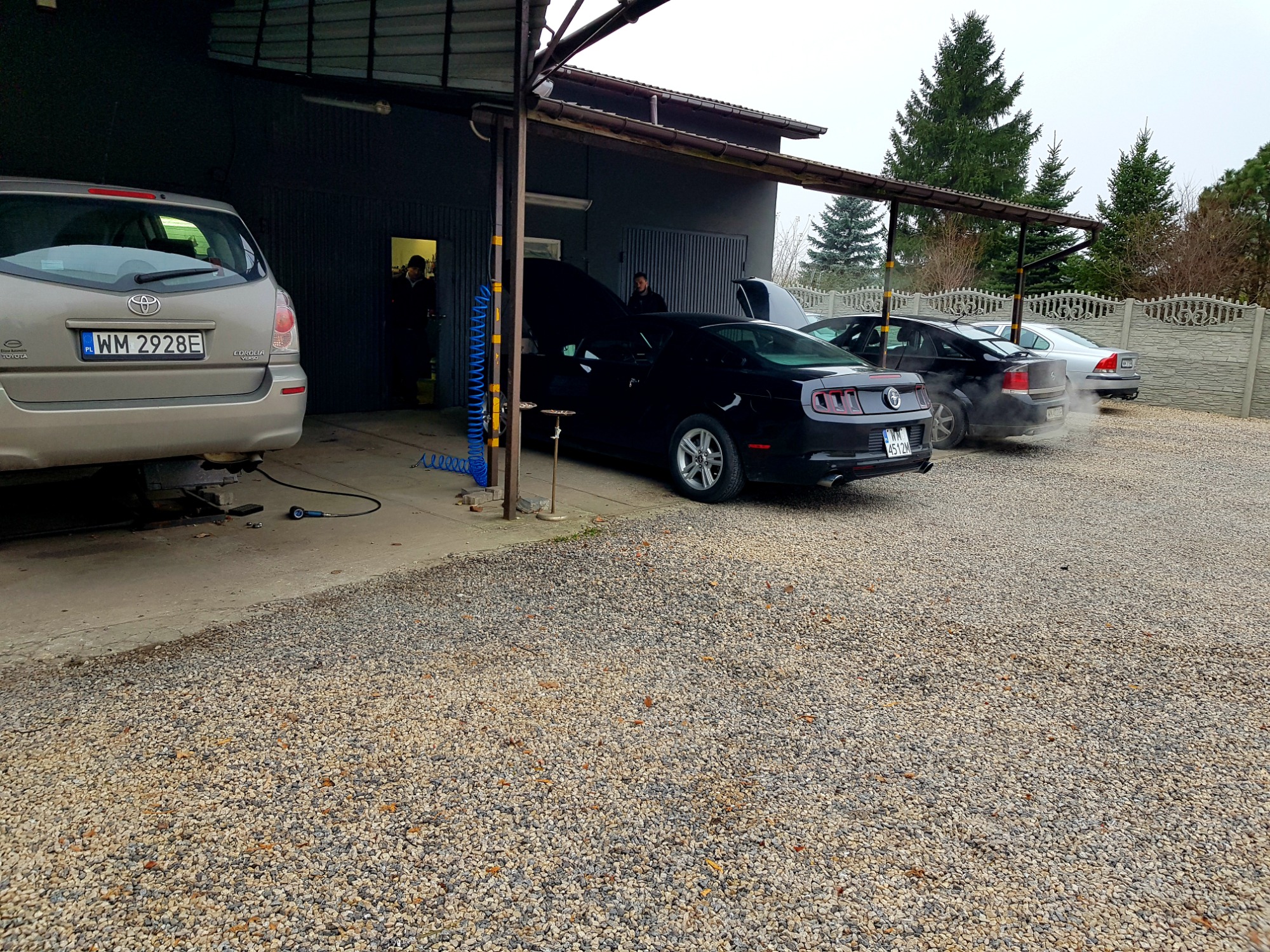 Warsztat samochodowy z kilkoma samochodami, w tym czarny Ford Mustang z otwartą maską, stojący na żwirowym parkingu przed budynkiem z otwartymi wrotami, gdzie widoczny jest mechanik.