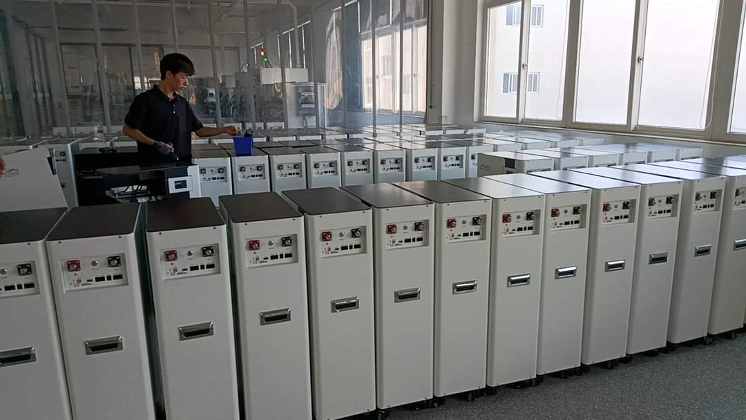 Rząd identycznych, białych szaf magazynów energii w hali produkcyjnej, pracownik w czarnym uniformie wykonuje prace montażowe przy jednym z urządzeń.