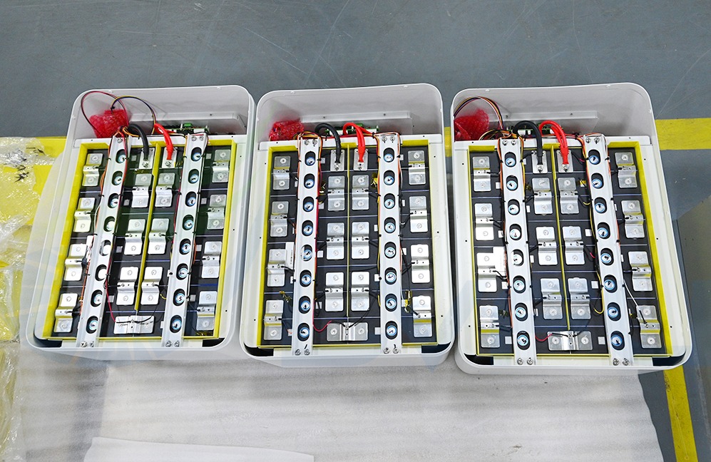 Trzy białe obudowy magazynów energii z żółtymi ramkami, widoczne wewnętrzne komponenty i okablowanie, zabezpieczone czerwonymi osłonami transportowymi.