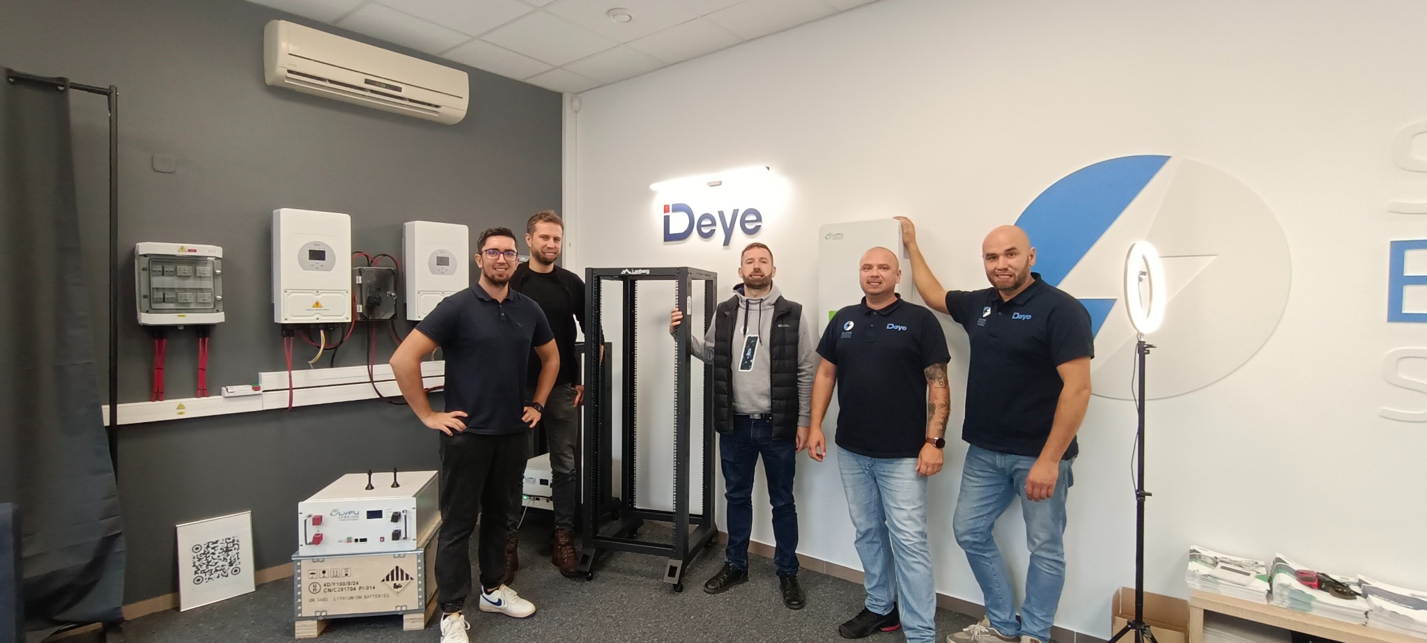 Grupa pięciu mężczyzn w pomieszczeniu z urządzeniami fotowoltaicznymi i magazynem energii, w tle inwertery, rozdzielnica i moduł bateryjny LVFU na palecie, na ścianie logo firmy Deye.