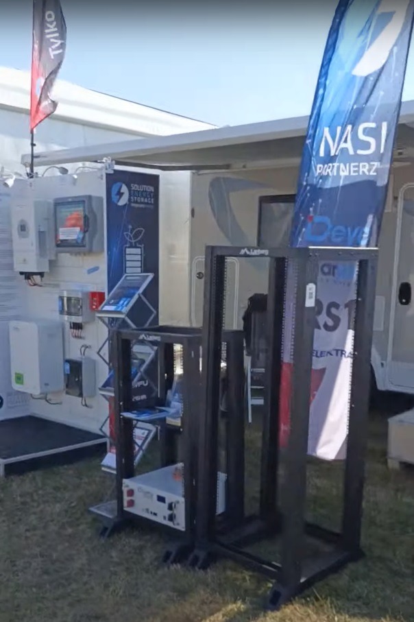 Ekspozycja zewnętrzna z modułami fotowoltaicznymi, inwerterami i magazynem energii na stoisku targowym z flagami reklamowymi.