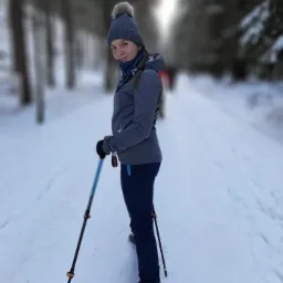 Kobieta w szarej kurtce i czapce z pomponem, spacerująca po ośnieżonej ścieżce w lesie, używająca kijków trekkingowych z niebieskimi elementami.