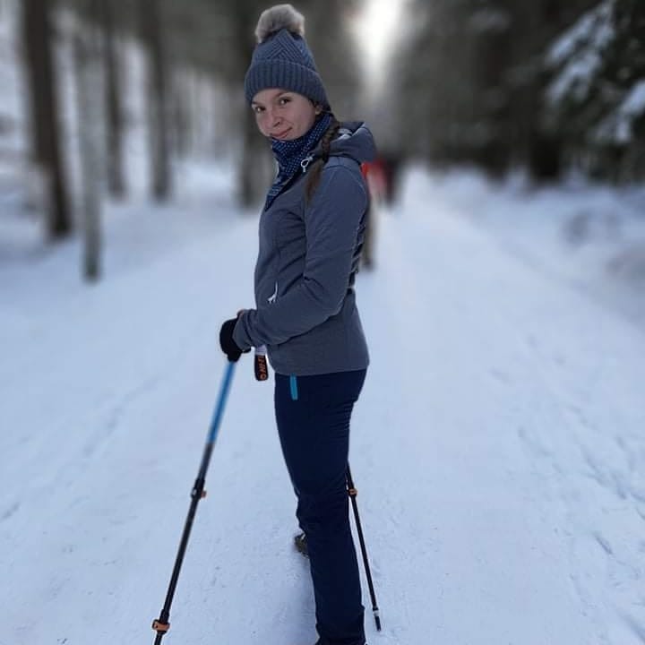 Kobieta w szarej kurtce i czapce z pomponem, spacerująca po ośnieżonej ścieżce w lesie, używająca kijków trekkingowych z niebieskimi elementami.