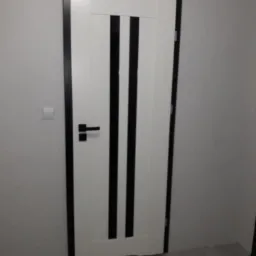 Białe drzwi wewnętrzne z czarnymi pionowymi paskami i czarną ramą, zamontowane w szarej ścianie, widoczny fragment podłogi.