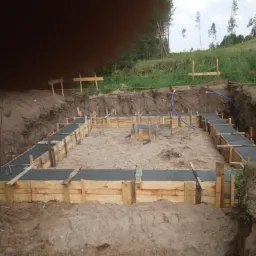 Fundament domu w trakcie budowy: drewniany szalunek wypełniony świeżym betonem, widoczne wystające pręty zbrojeniowe, ziemny wykop, w tle trawa i drzewa.