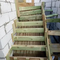 Wewnętrzne schody betonowe w trakcie budowy, z drewnianym szalunkiem i zbrojeniem stalowym, widoczne białe ściany z betonu komórkowego.