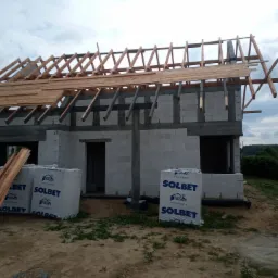 Budowa domu z betonu komórkowego w trakcie wznoszenia dachu, widoczne krokwie i częściowe poszycie, na ziemi paczki bloczków SOLBET