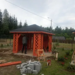 Montaż pomarańczowej altany z czarnym dachem w ogrodzie, widoczny pracownik z miotłą, narzędzia i kostka brukowa.