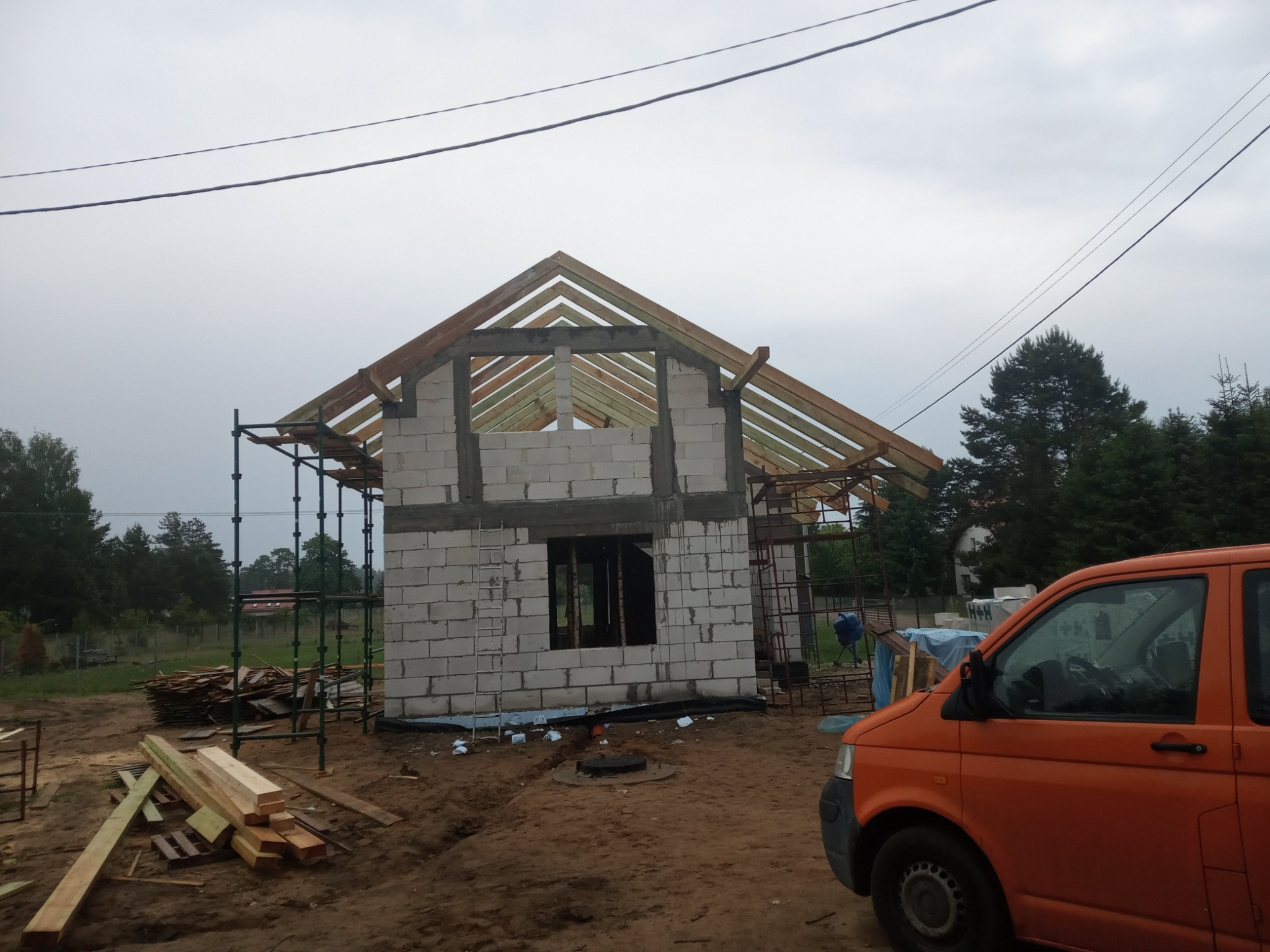 Wznoszona konstrukcja domu z bloczków betonowych, widoczne drewniane elementy więźby dachowej i rusztowania, na pierwszym planie pomarańczowy samochód dostawczy i stos drewna.