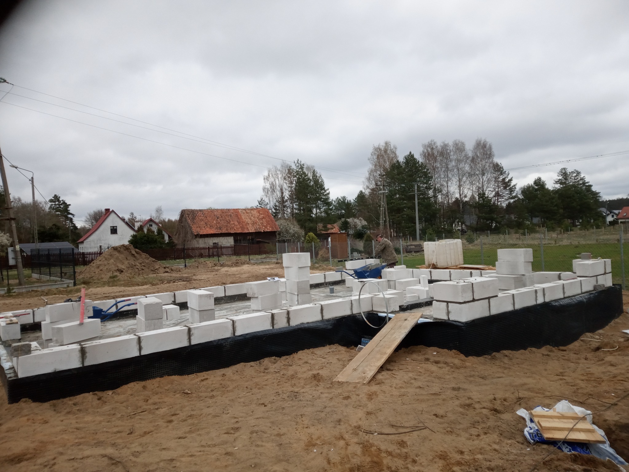 Budowa fundamentów z bloczków betonowych na czarnej folii izolacyjnej, widoczny krajobraz wiejski z domami i drzewami w tle, pochmurne niebo.