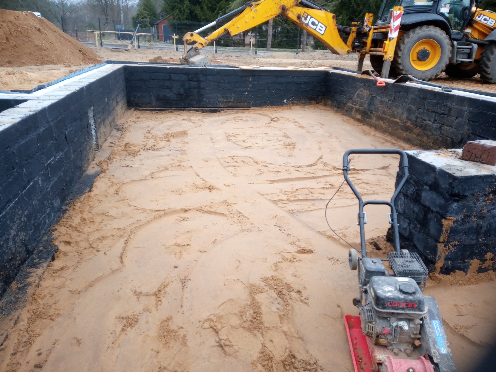 Wykop pod fundamenty z izolacją przeciwwilgociową, ubijarka wibracyjna i koparka JCB w tle.