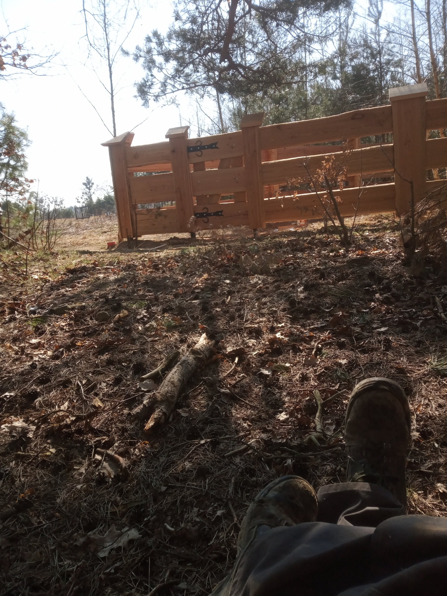 Niedokończone, surowe drewniane ogrodzenie z bramką w lesie, widok z dołu na buty osoby siedzącej na ziemi, z suchymi liśćmi i gałęziami.