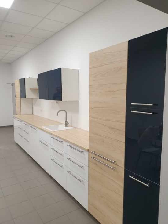 Nowoczesna zabudowa kuchenna z białymi szafkami dolnymi z długimi, chromowanymi uchwytami, drewnopodobnymi blatami i wysokimi szafkami w kolorze jasnego drewna i czarnego połysku, widoczna biała...