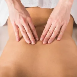 Dłonie masujące plecy kobiety leżącej na białym ręczniku, dekoracja z kwiatów po bokach.