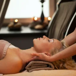 Kobieta z blond włosami relaksuje się podczas masażu szyi i karku w gabinecie, w tle świece i zasłony.