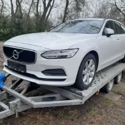 Białe Volvo kombi na metalowej przyczepie samochodowej z niebieskim pokrowcem zabezpieczającym na haku holowniczym, tło park z drzewami.