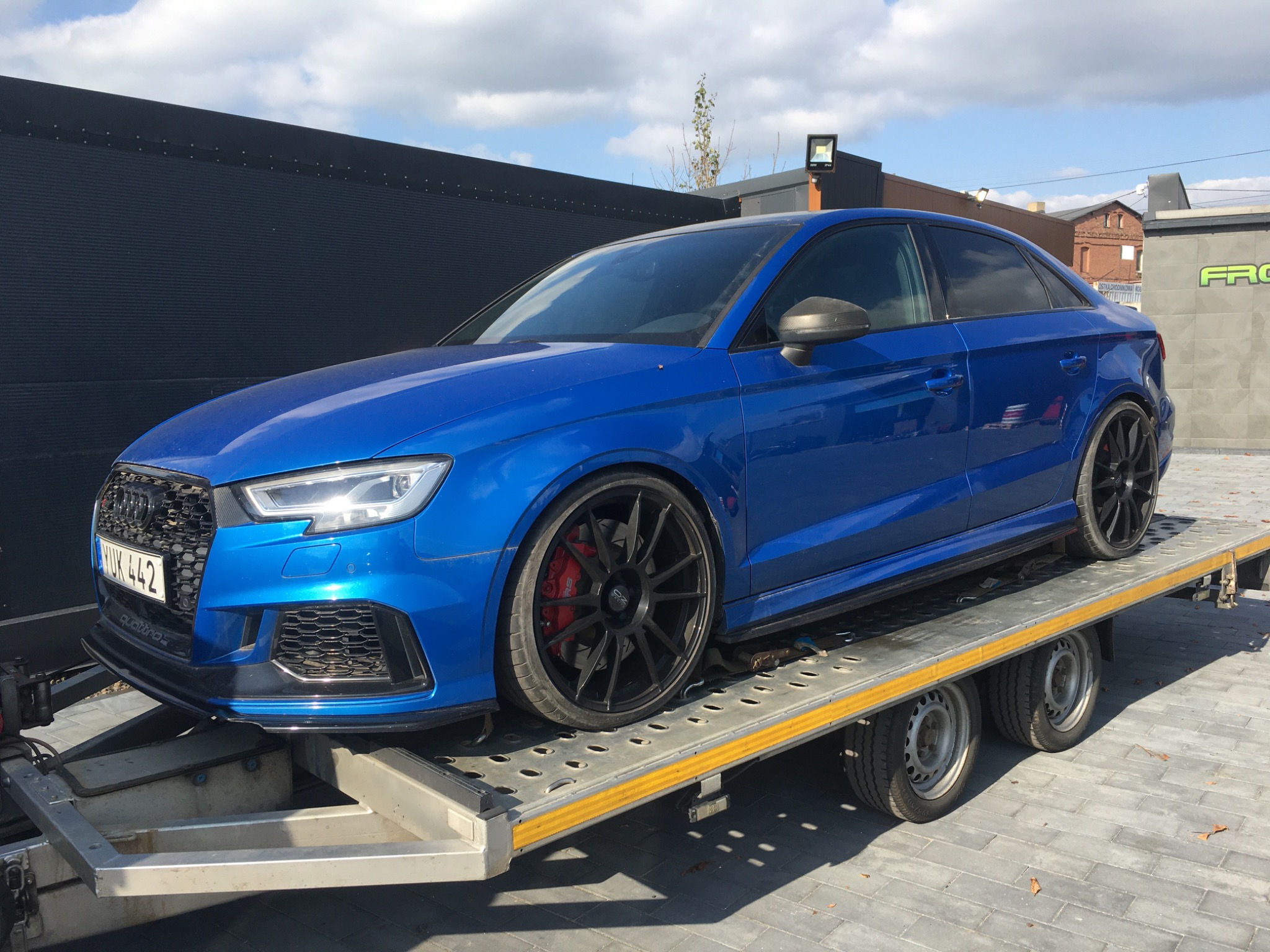 Niebieski samochód osobowy marki Audi z czarnymi felgami, umieszczony na platformie transportowej z dwoma osiami, widoczny na tle czarnego budynku.