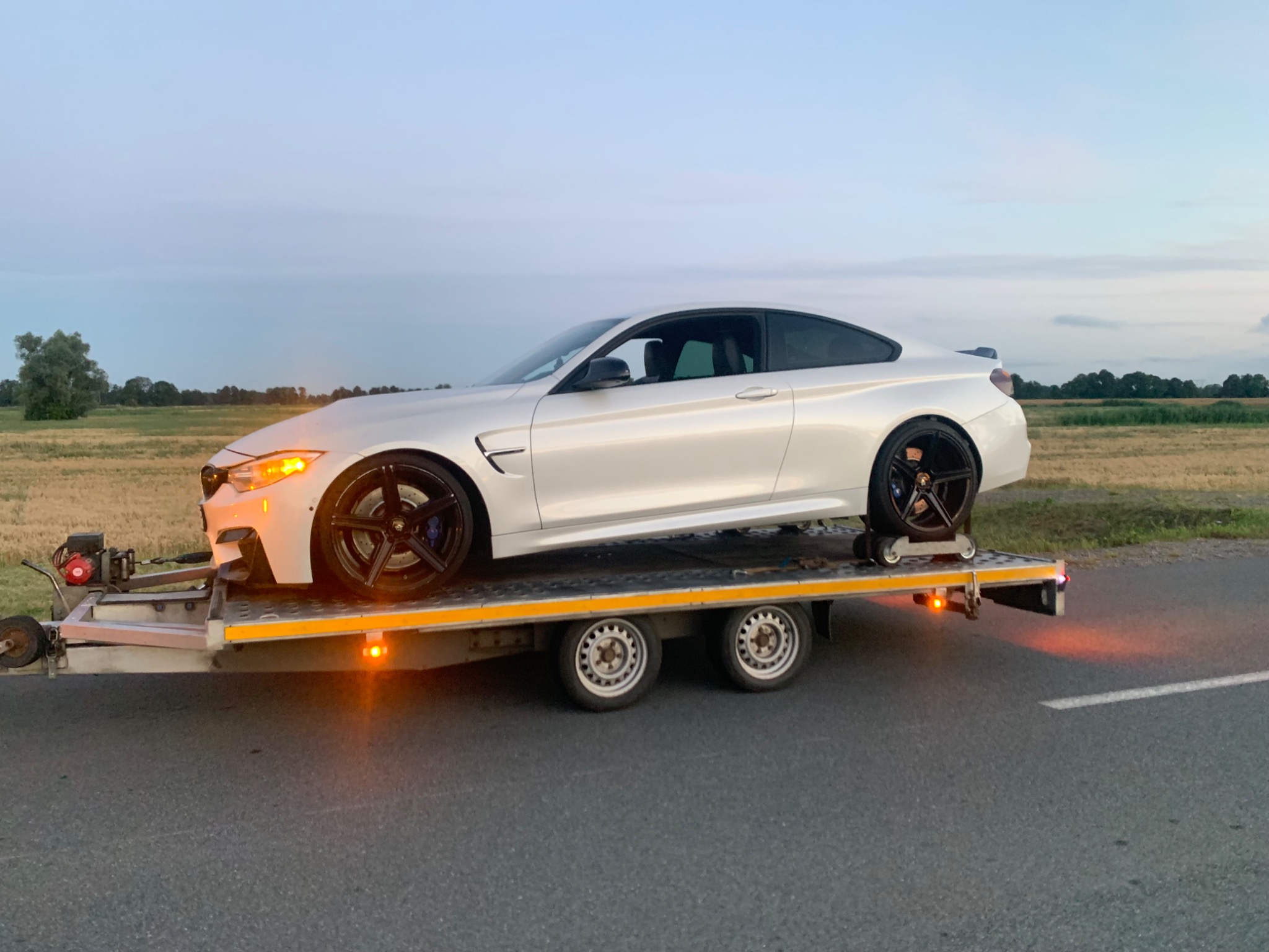 Białe sportowe auto na niskopodwoziowej przyczepie w trakcie transportu, widoczne pasy zabezpieczające koła, w tle pole i zachodzące słońce.