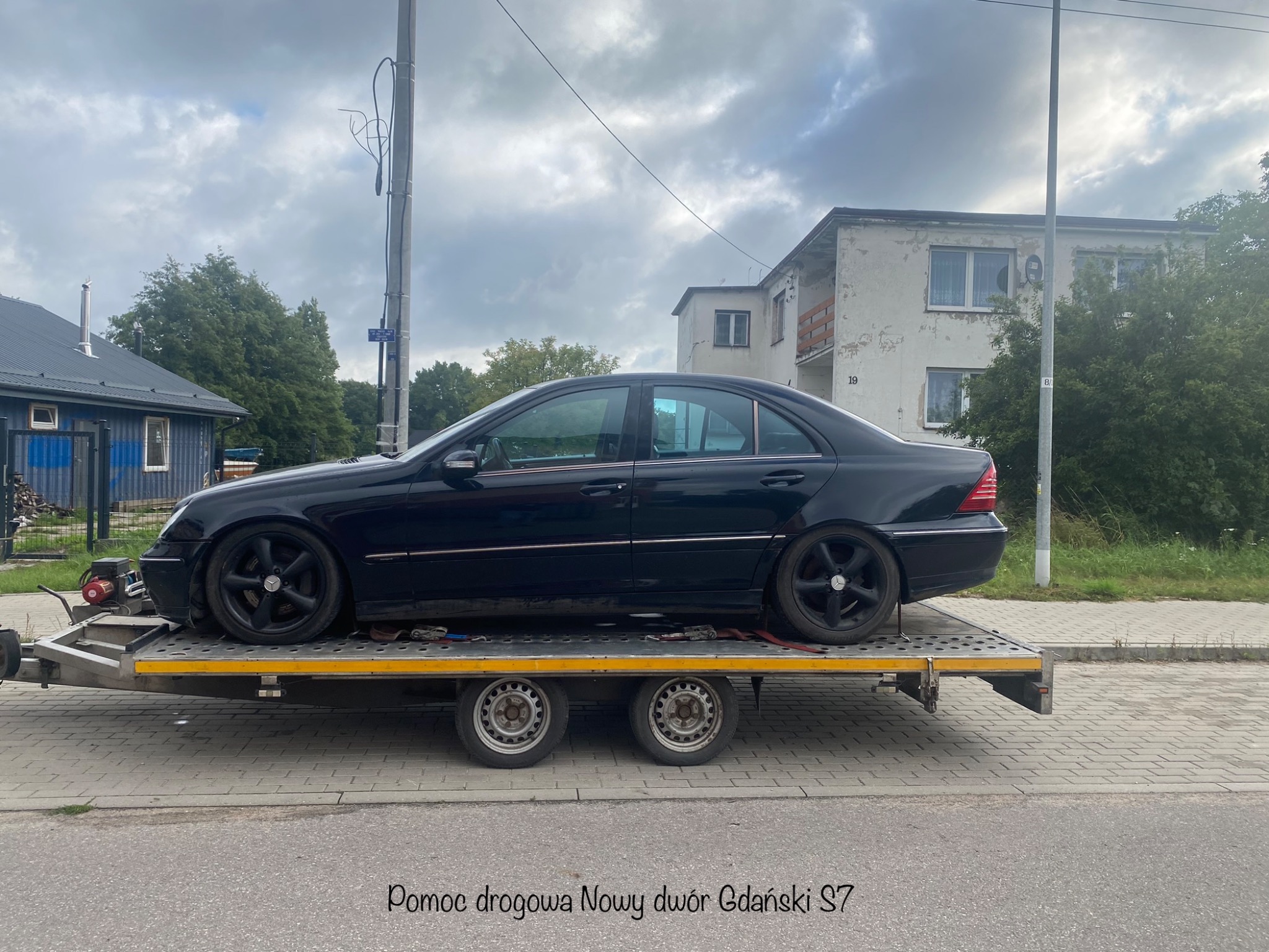 Czarny Mercedes-Benz klasy C z czarnymi felgami transportowany na niskopodwoziowej przyczepie na tle budynku mieszkalnego i pochmurnego nieba.