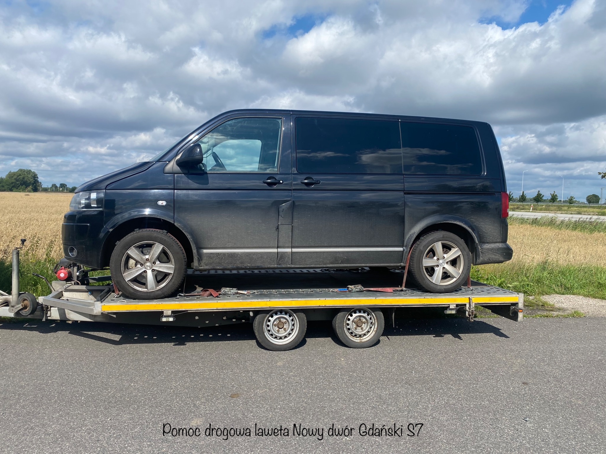 Czarny van przewożony na platformie transportowej z żółtymi pasami mocującymi, na tle pola i pochmurnego nieba.