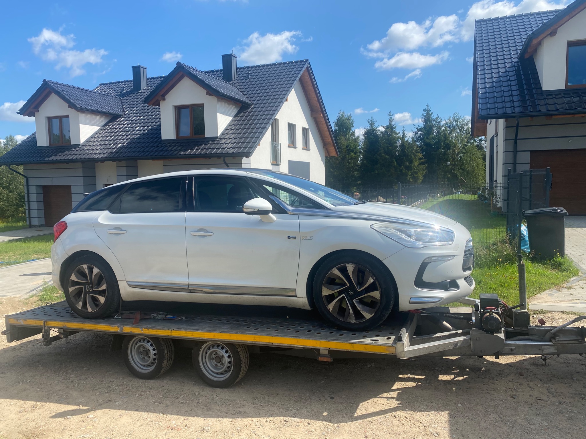 Biały samochód osobowy marki Citroen DS5 na dwuosiowej przyczepie samochodowej z żółtymi elementami, na tle dwóch domów jednorodzinnych w słoneczny dzień.