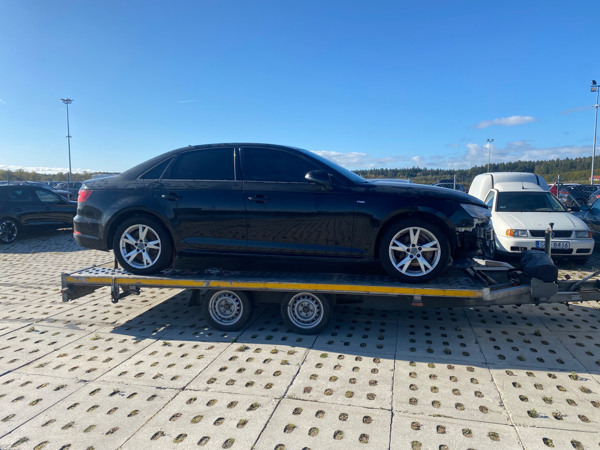 Czarny samochód osobowy Audi A4 bez przedniego zderzaka, przewożony na żółtej lawecie na parkingu z betonową kostką, błękitne niebo w tle.