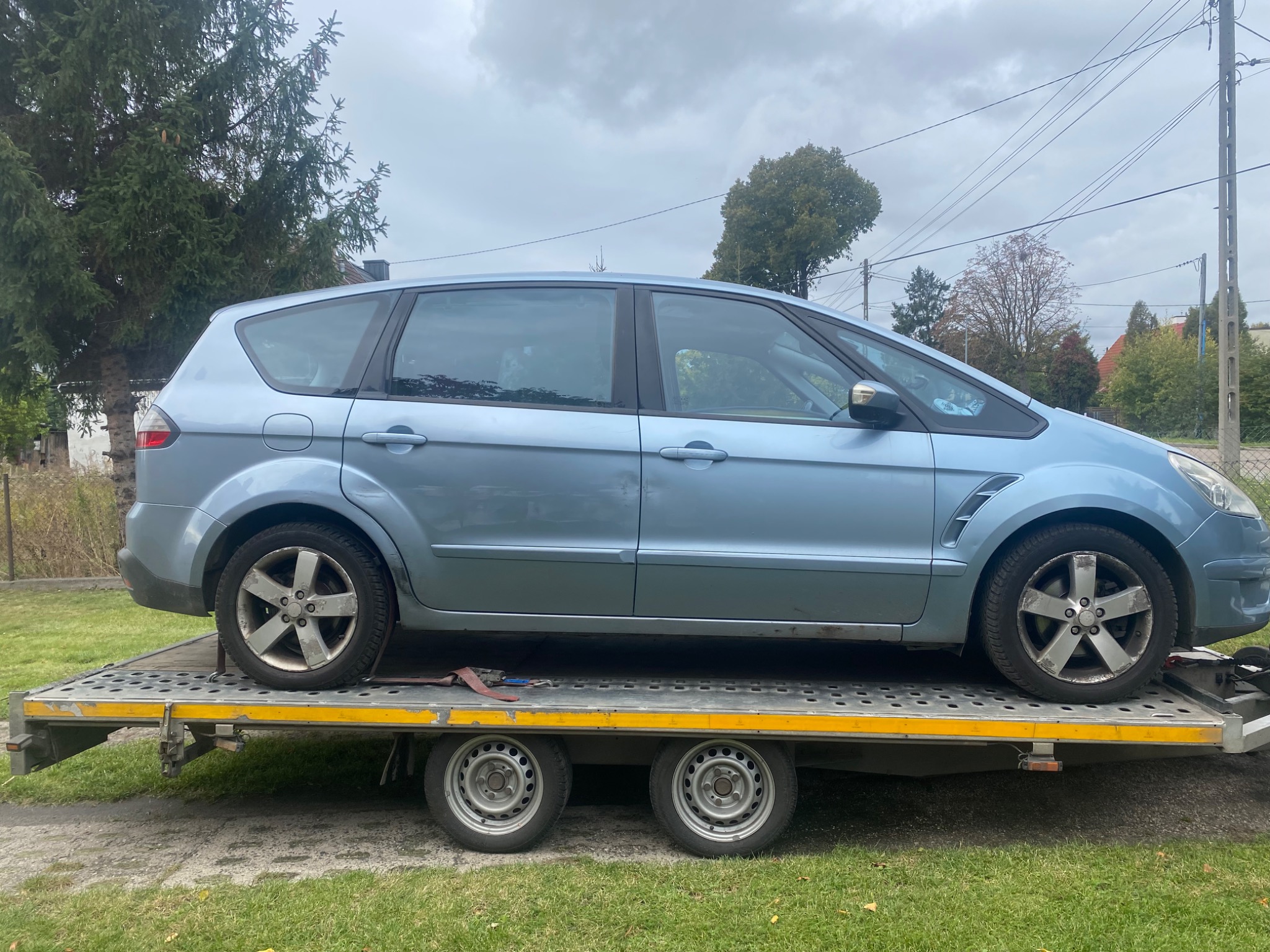 Niebieski samochód osobowy typu minivan zabezpieczony pasami transportowymi na przyczepie samochodowej z metalową kratownicą, widok z boku na tle zielonego trawnika i drzew