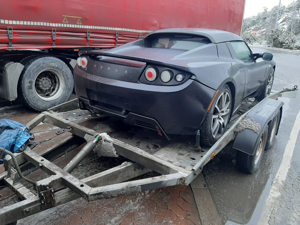 Czarny samochód sportowy Tesla Roadster zabezpieczony na metalowej platformie przyczepy transportowej, częściowo widoczna czerwona naczepa ciężarówki w tle.